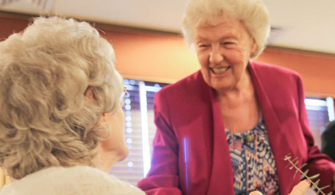 An elderly woman in a pink blazer talking to another elderly woman